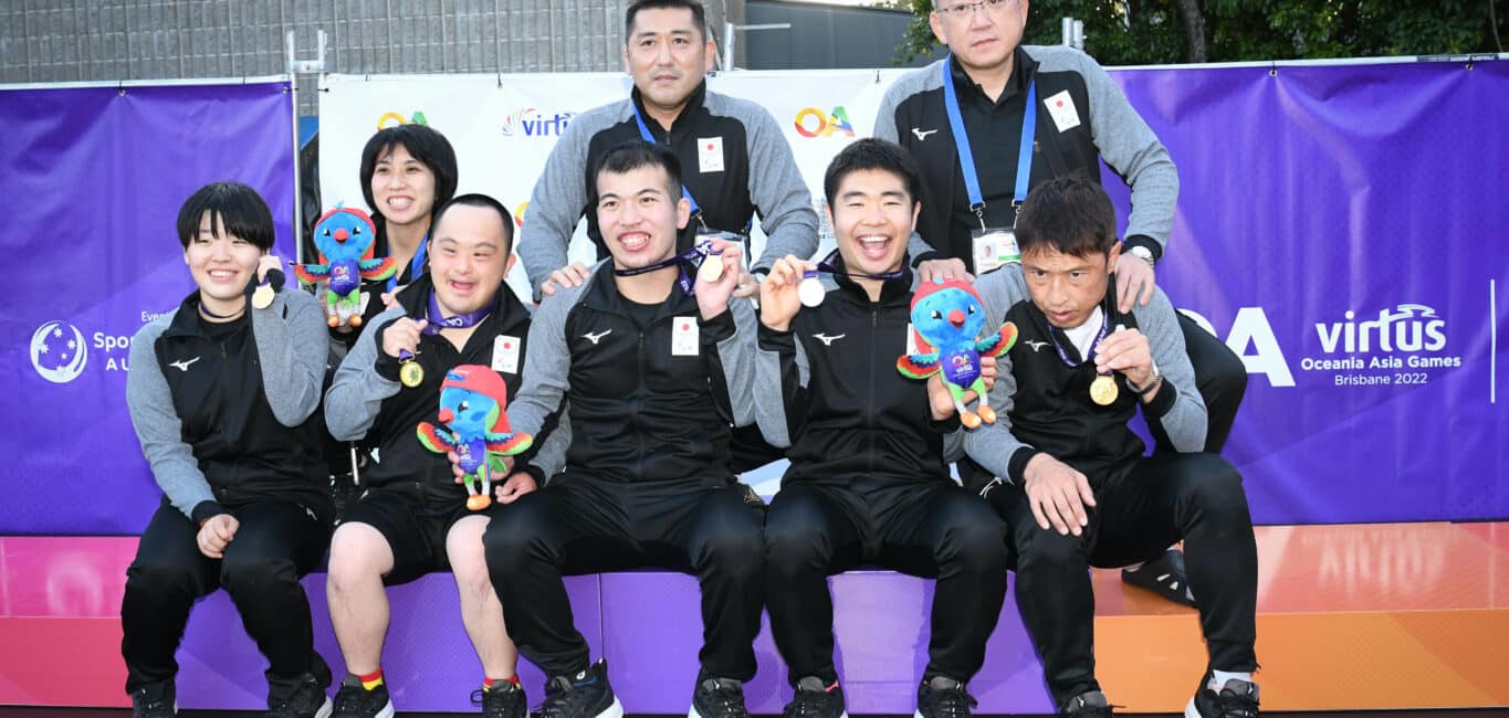 Six Japanese sitting on the podium, with two coaches behind them, holding up their medals that are around their necks, and Lorri plushes.
